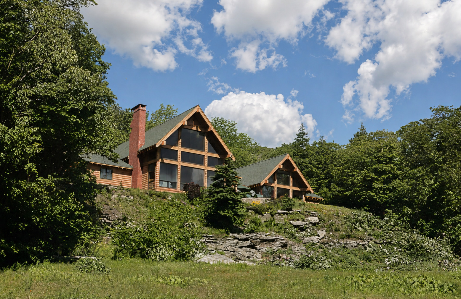 The house from the meadow below: a log and timber lodge set into a wooded hillside, its tall gable end filled with a grid of glazing, a second smaller glazed gable beyond it, and a brick chimney rising past the roof.