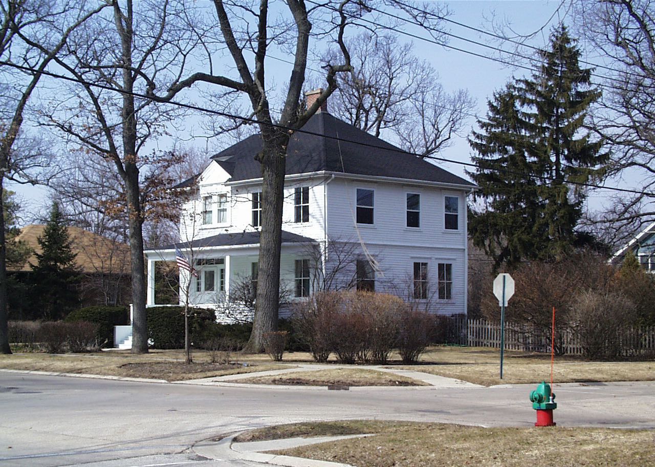 The house before the work: a white two-storey house on a street corner, its porch stopping at the front elevation and the side wall meeting the garden plainly.