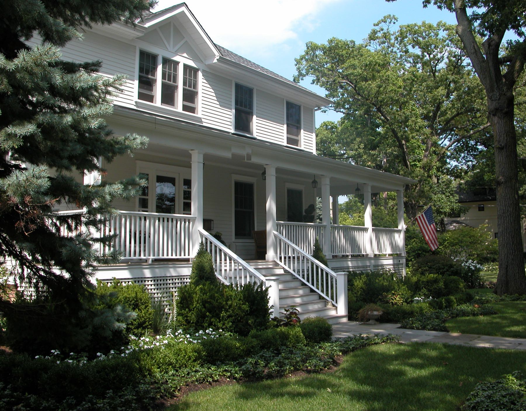 The front of a white clapboard house behind a deep porch with turned balusters, reached by a broad flight of steps through mature planting.