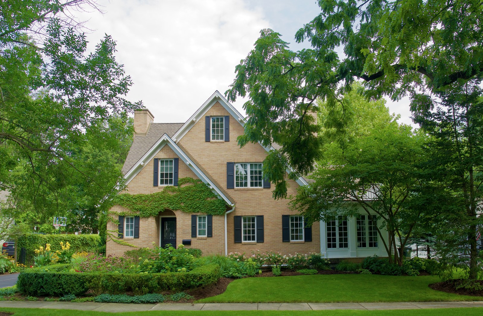 The full street elevation, with two steep gables side by side, dark shutters at every window, ivy swagged across the arched entry, and the sun room projecting at the right-hand end.
