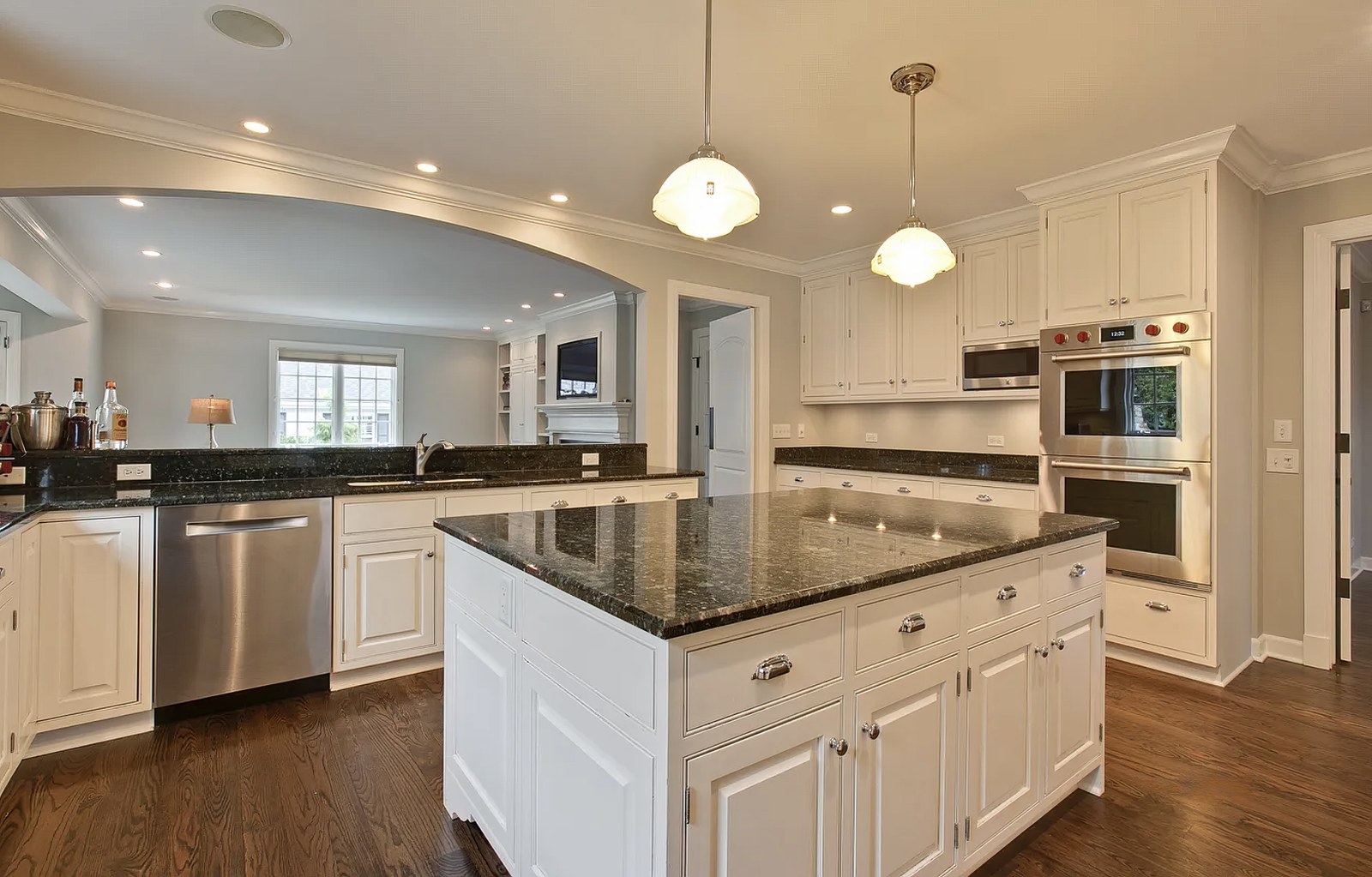The kitchen from the sink end, showing the island, the double wall ovens set into the cabinetry, and the arch opening to the living room beyond.