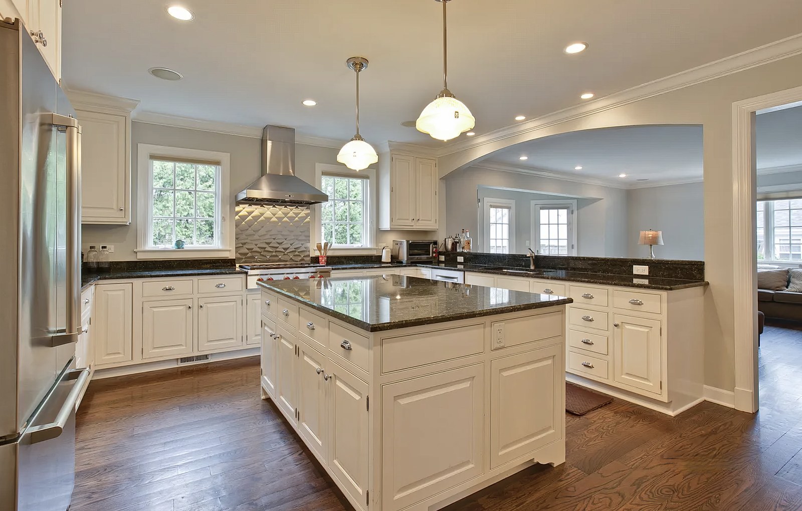 The kitchen, with a dark granite island under two schoolhouse pendants, cream cabinetry, a stainless hood between two windows, and an arched opening through to the living room.