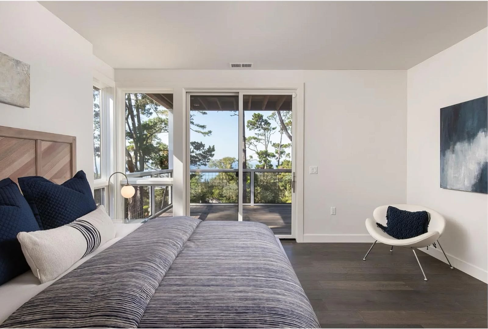 Lower level bedroom with a chevron timber headboard, sliding doors onto a railed deck and a glimpse of ocean through the pines.