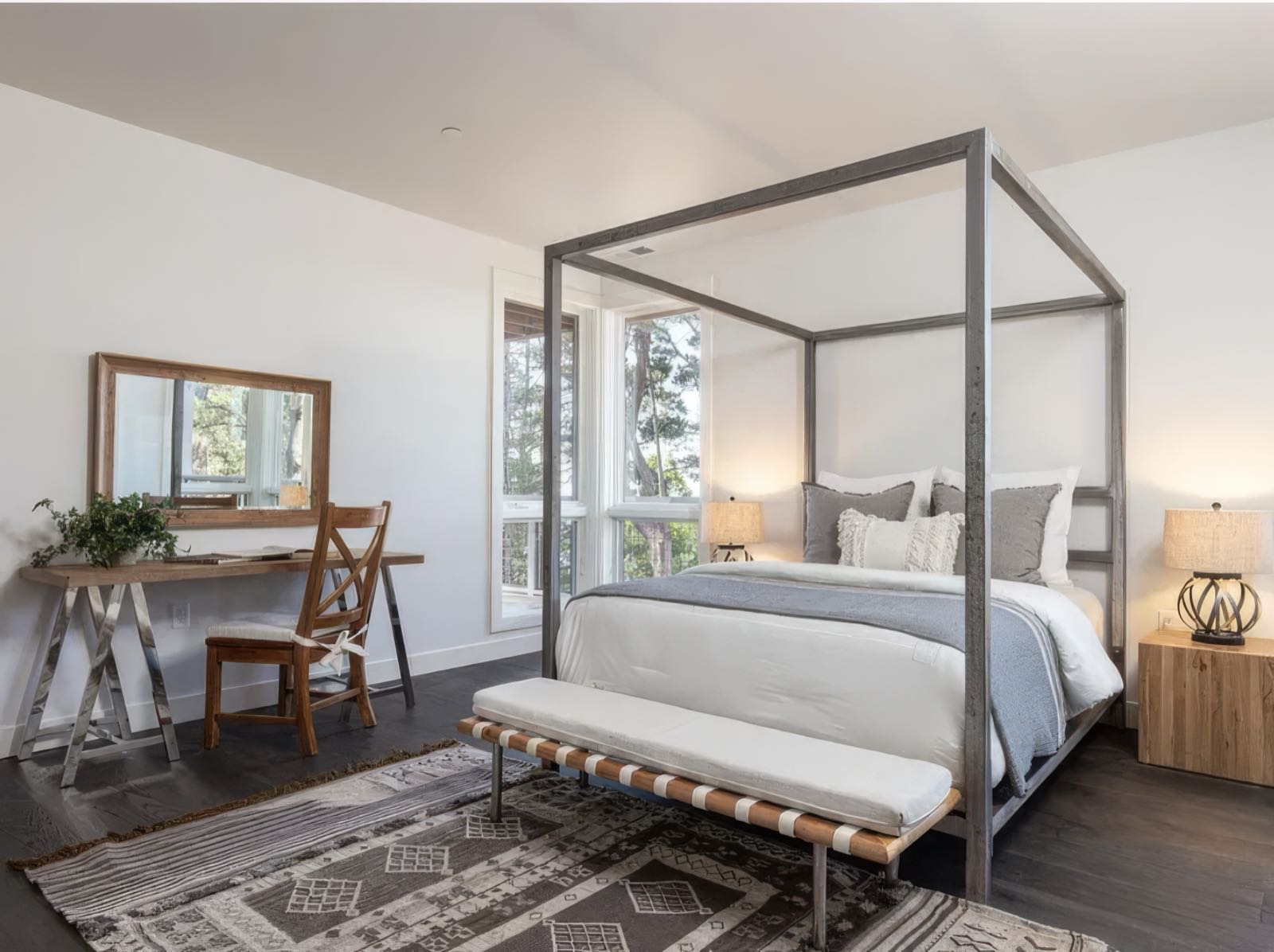 Lower level bedroom with a steel four-poster frame, a sawhorse desk and a window onto the trees.