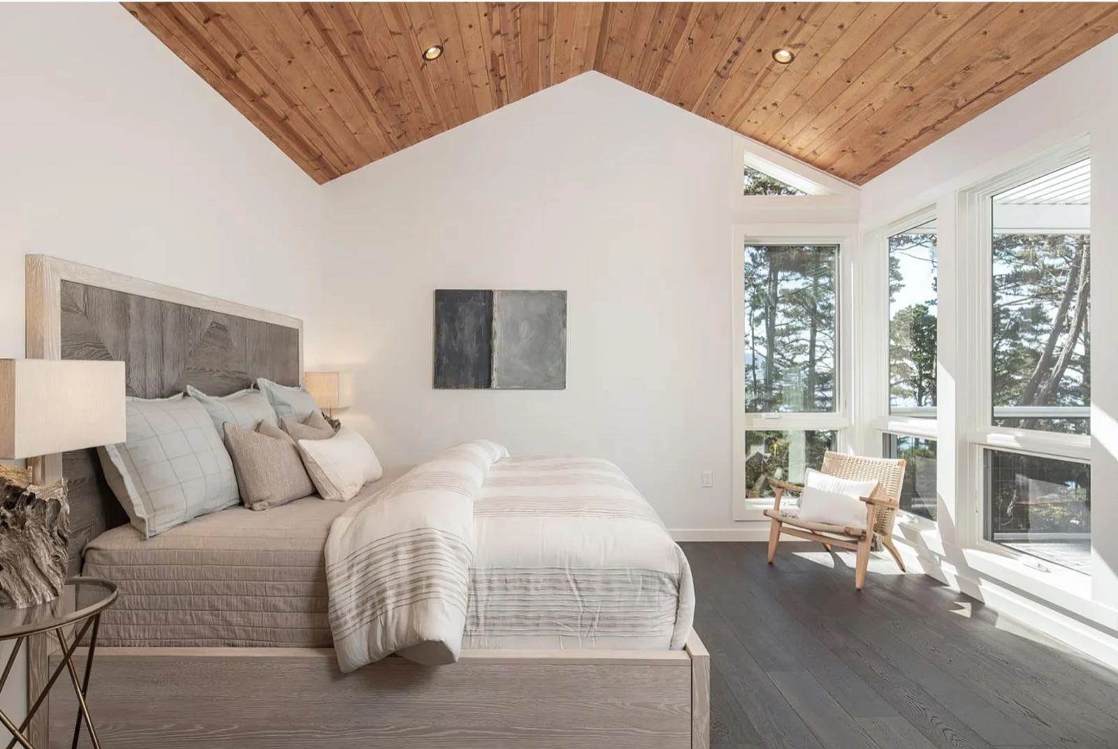Primary bedroom under a vaulted timber ceiling, with corner windows and a low chair in the light.