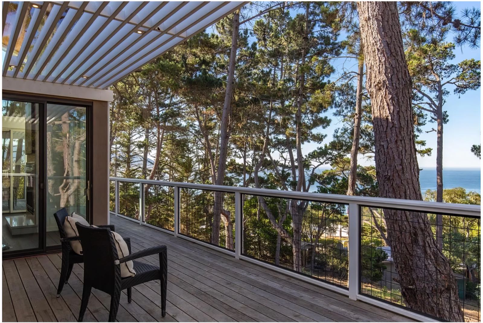 Deck with a slatted pergola overhead, cable railing, and a view out through the pines to the sea.