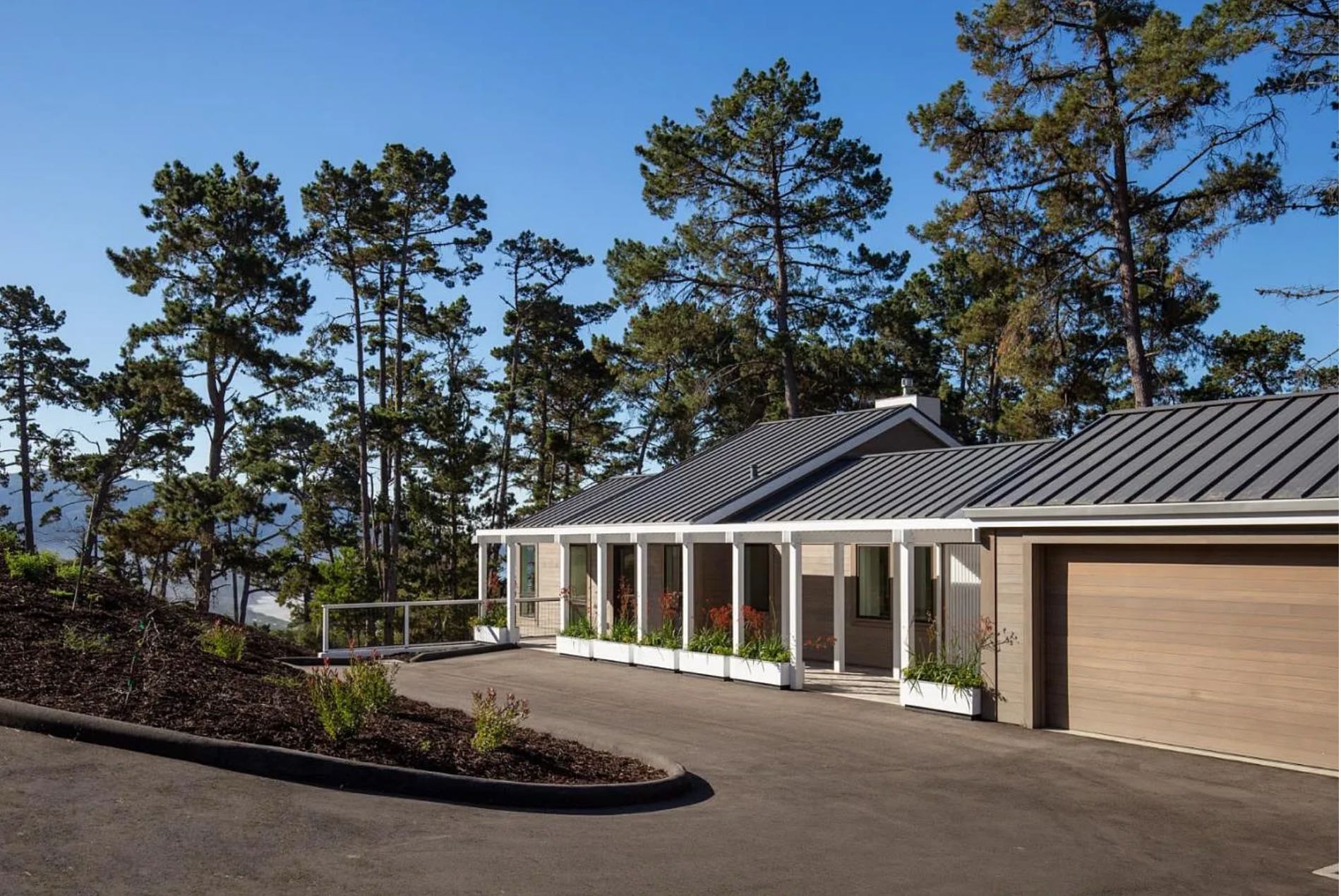 The front of the house from the drive: a colonnaded walk running the length of the glazed face, under a dark standing-seam roof, with a timber garage door at the right.