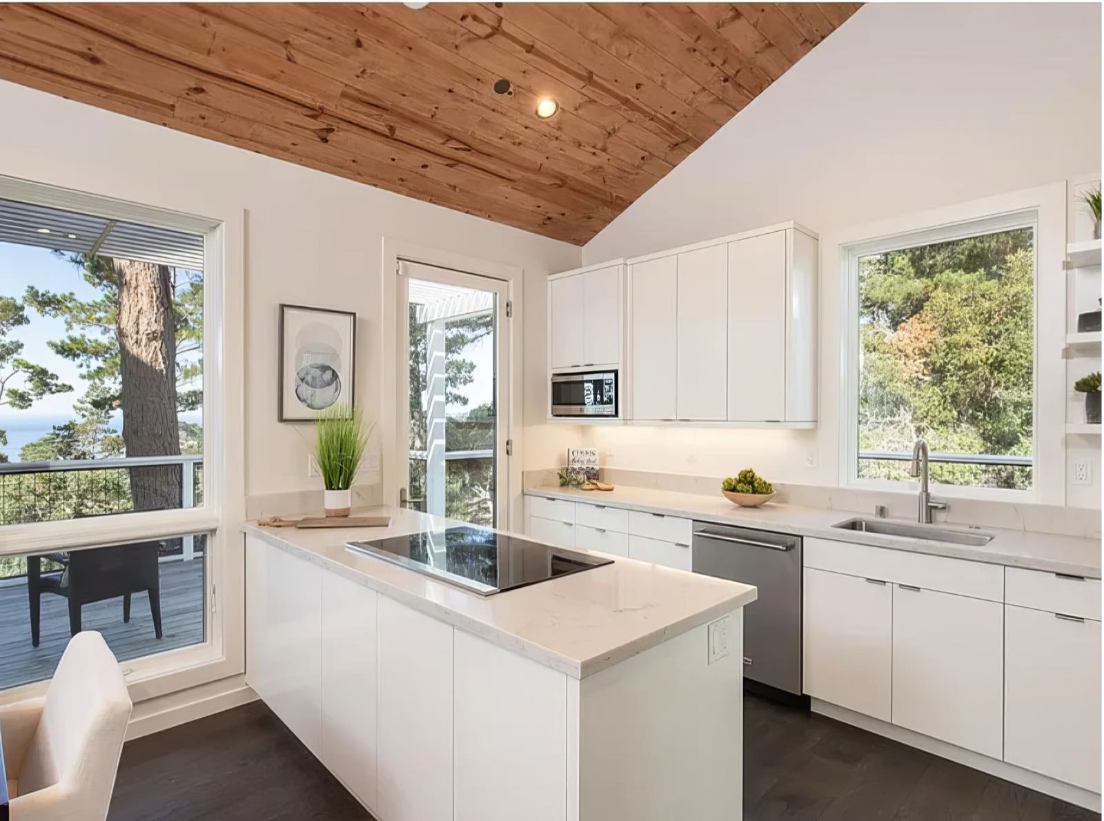 White kitchen with a quartz island and induction hob under a vaulted tongue-and-groove timber ceiling, windows on two sides.