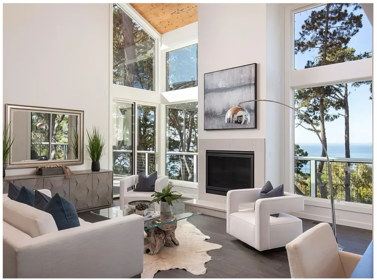 Double-height living room with a plaster fireplace, clerestory windows under a vaulted timber ceiling, and full-height glazing to the pines and ocean.