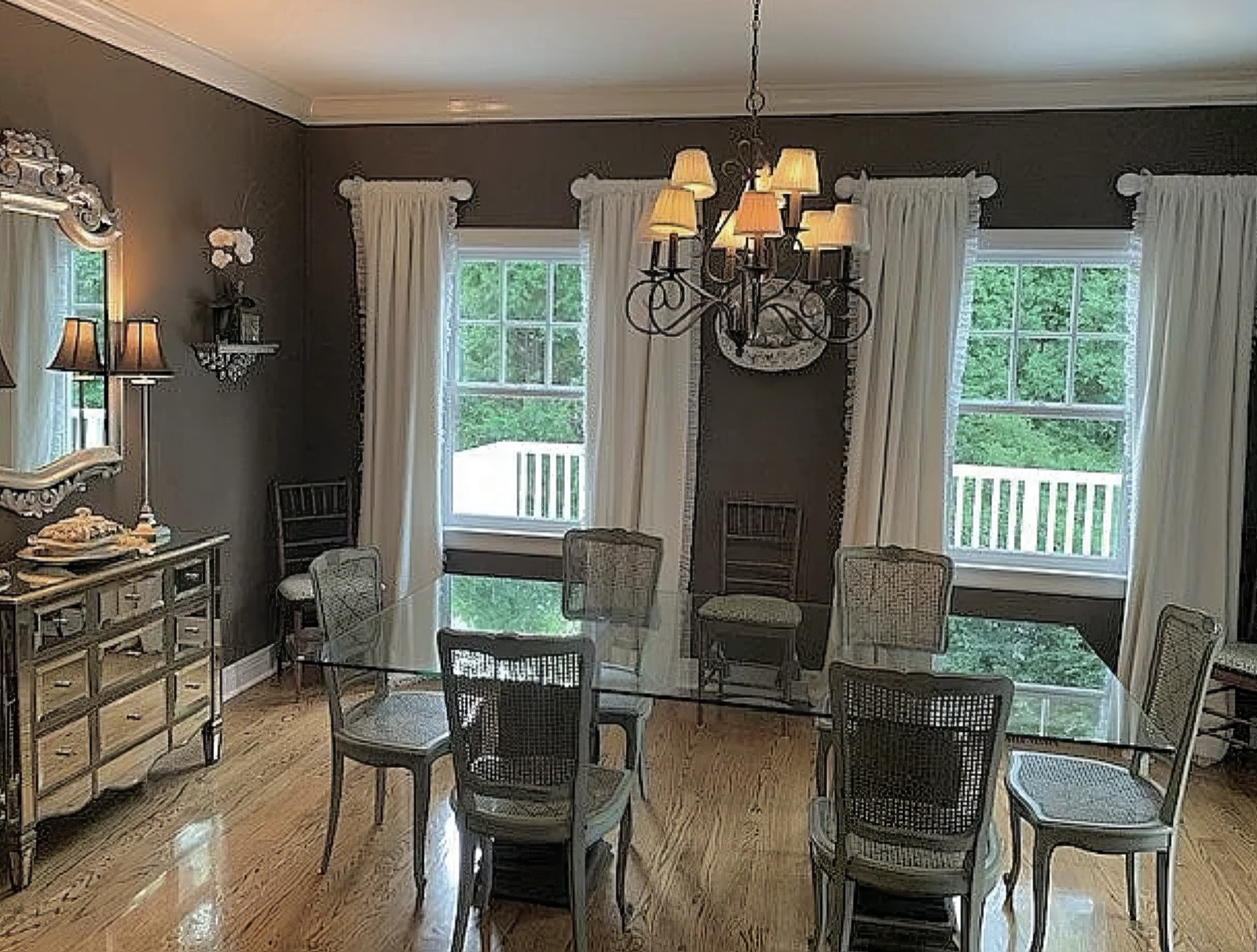 The dining room, its walls a deep brown against a white cornice and white curtains, with a glass-topped table, cane-backed chairs, an iron chandelier and three windows onto the deck.