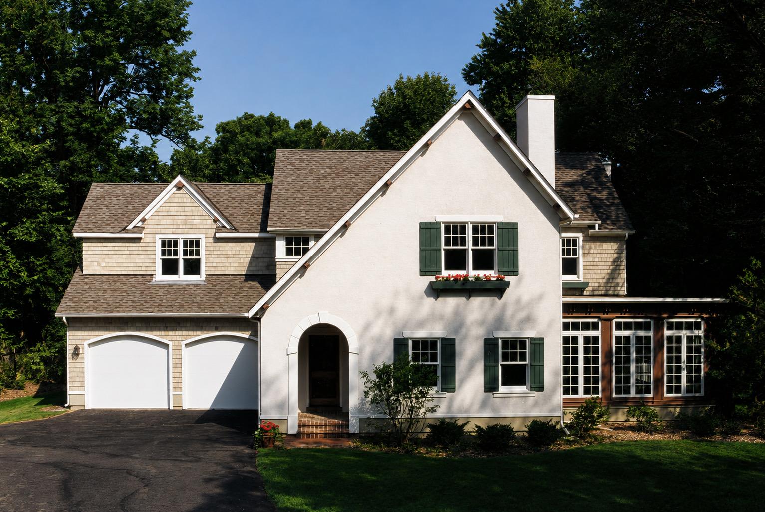 The house from the drive: a steep stuccoed gable with green shutters and a planted window box, a shingled garage wing stepping back to the left, and the glazed sun room under a low roof to the right.