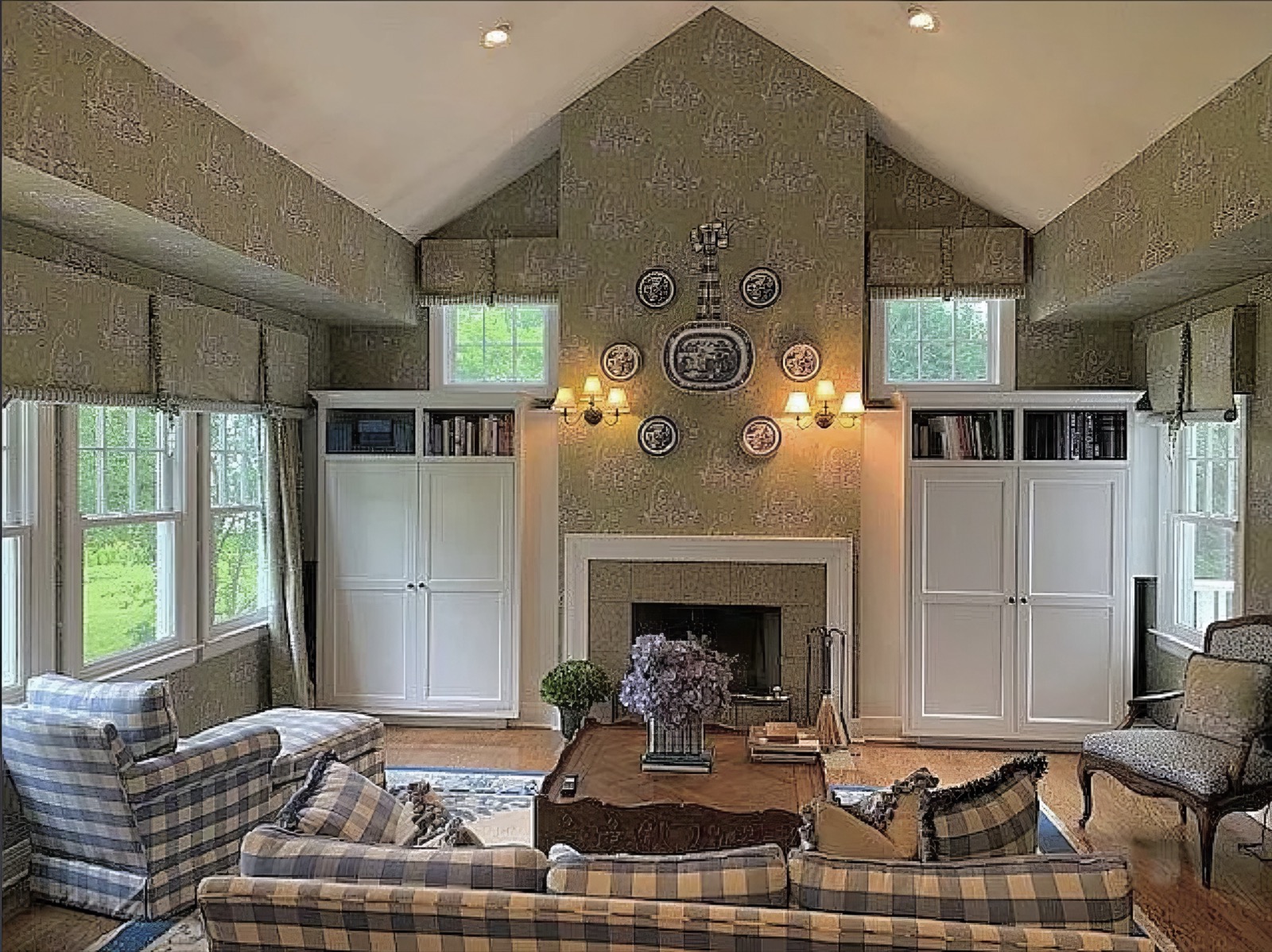 The family room under a ceiling vaulted to the gable, with a white-mantelled fireplace between two tall built-in cupboards, blue and white plates set around the chimney breast, and windows to either side.