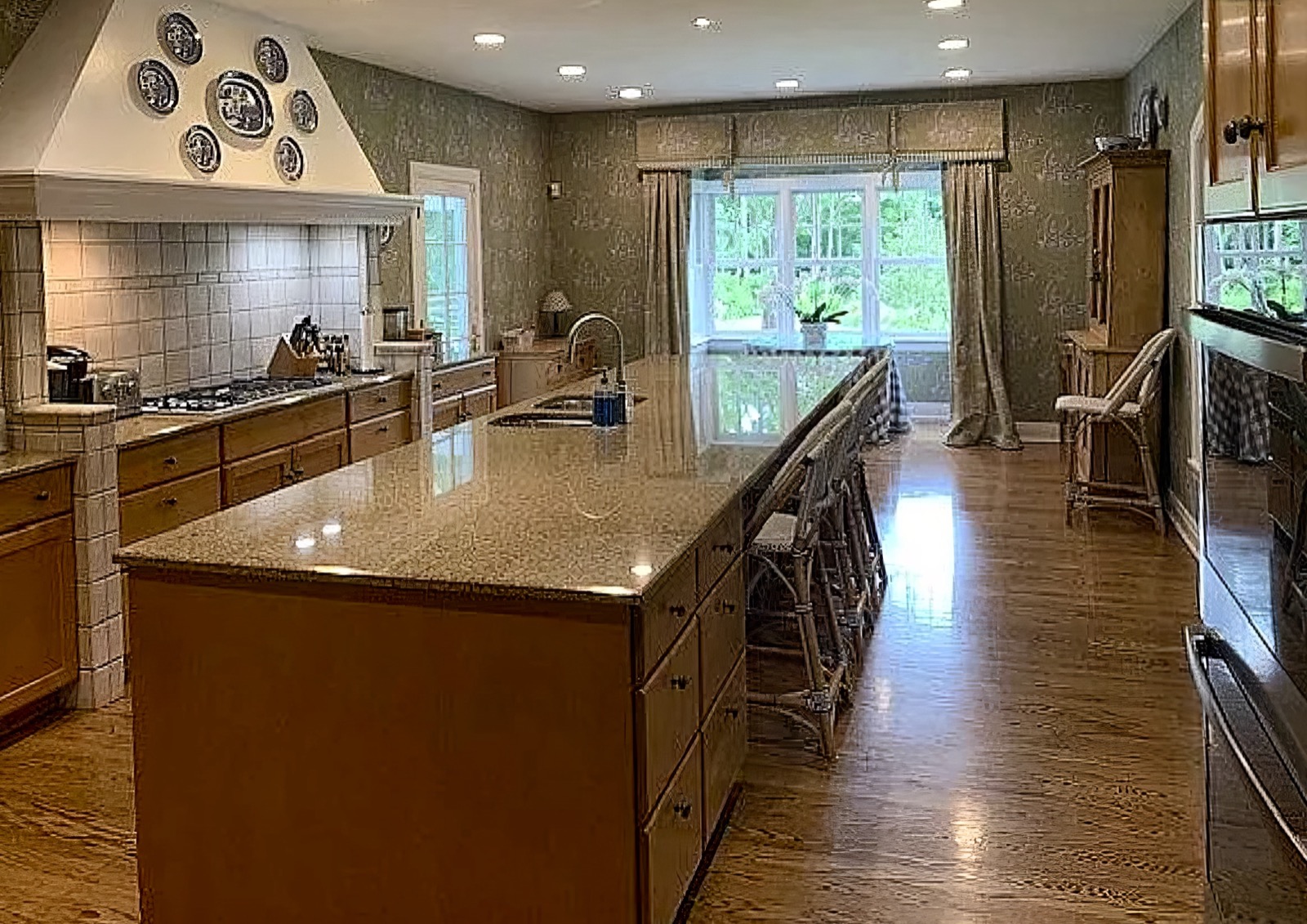 The kitchen along its length, with a long granite island and stools down one side, wood cabinetry, a white plaster hood hung with blue and white plates, and a curtained window at the far end.