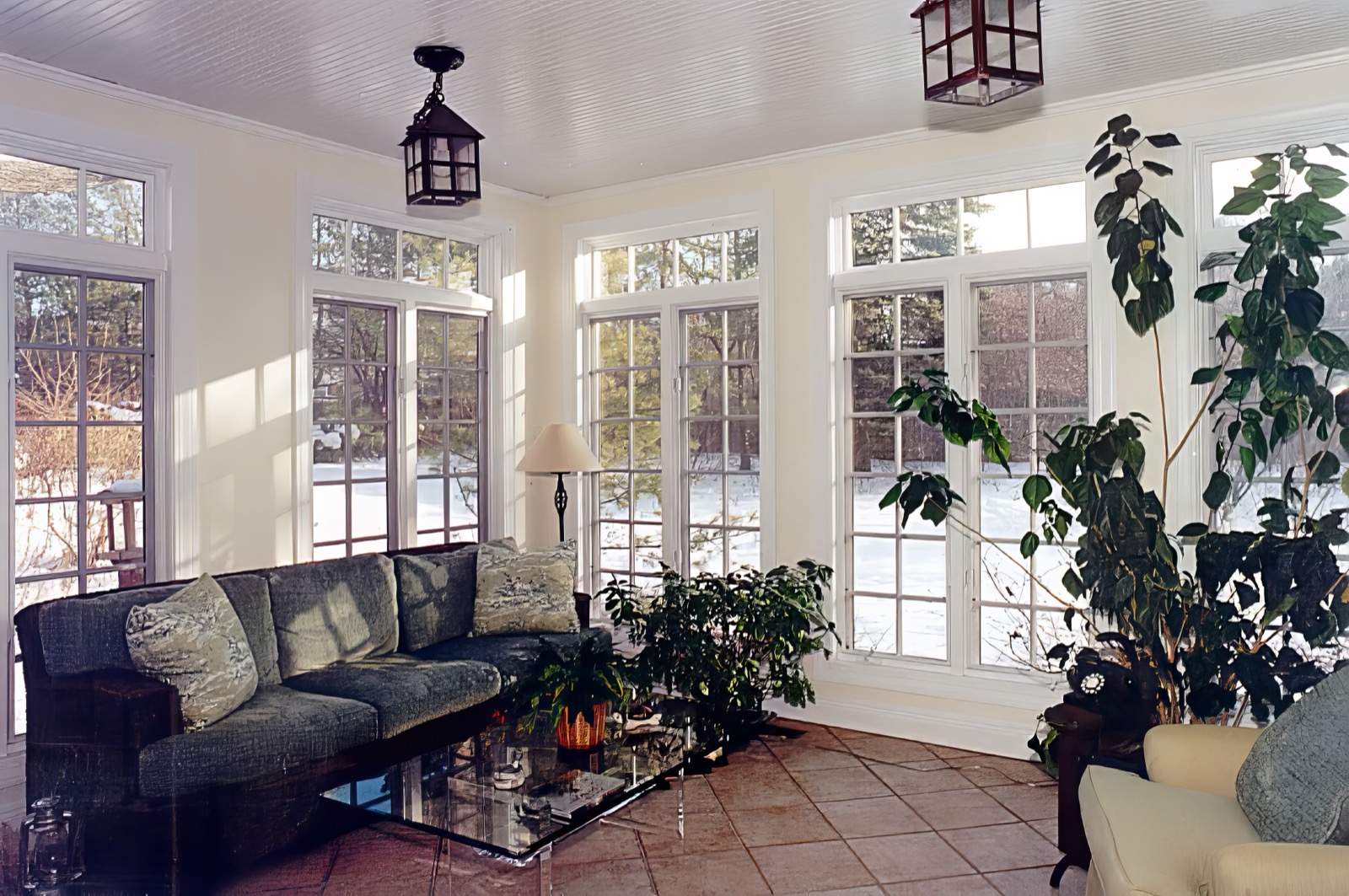 The sun room, glazed on three sides with divided-light windows and doors, a white beadboard ceiling carrying two lantern pendants, a tiled floor, and snow in the garden beyond.