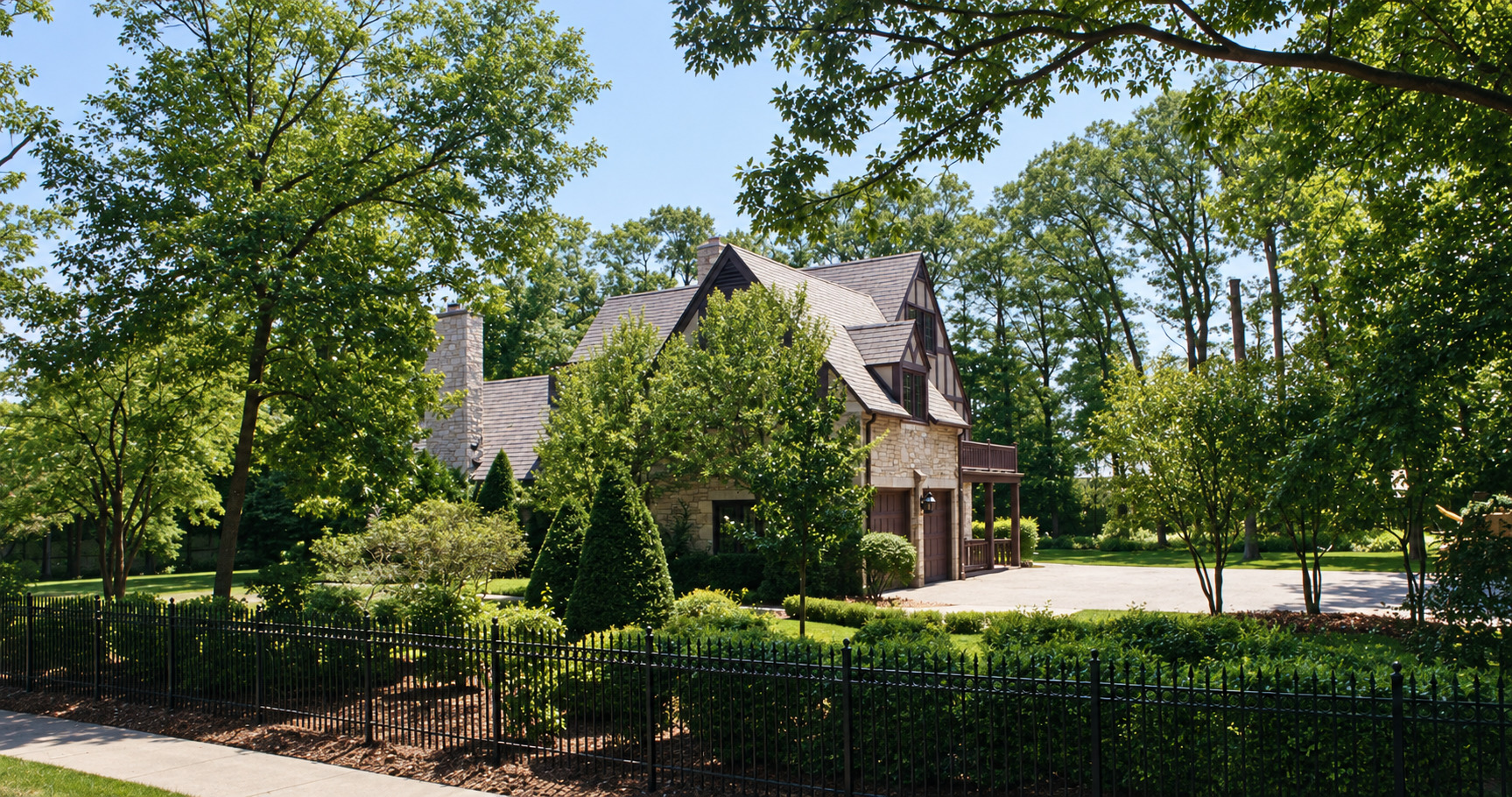 The house before the addition, from the same spot: the stone Tudor ending in a gable with a small balcony over a single garage door and a low porch beside it, trees close around.