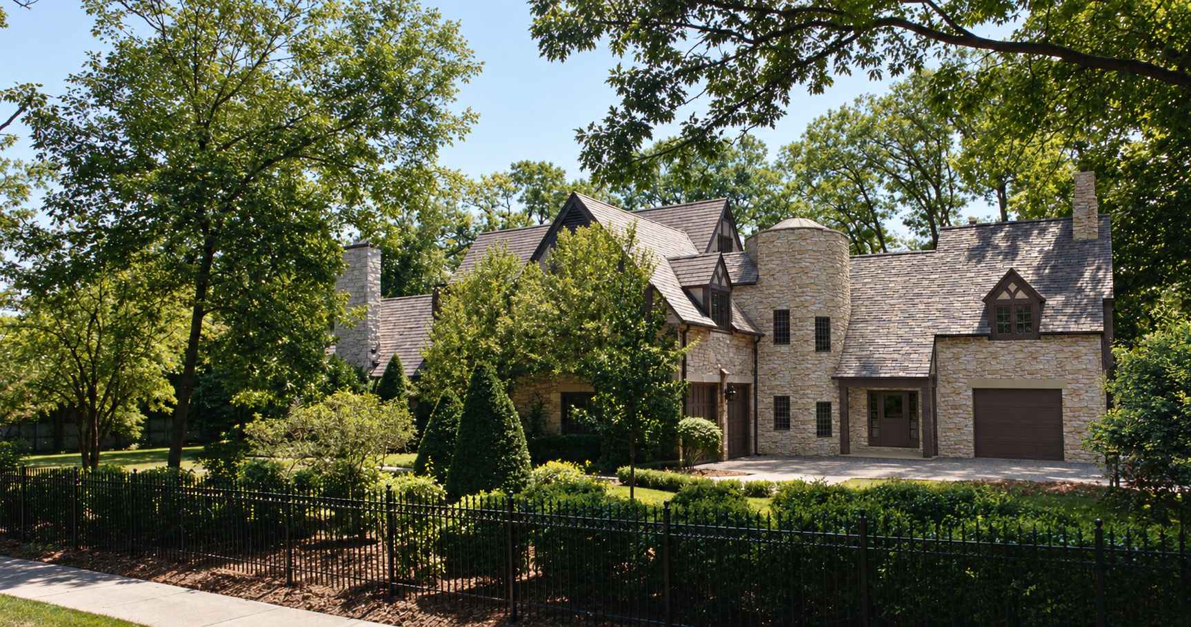 The house from the street after the addition: a stone Tudor with steep gables and timbered dormers, a round stone tower with a conical roof at the join, a brown-canopied entrance beside it, and a new gabled bay with a garage door at the far end, behind a wrought-iron fence and clipped hedges.