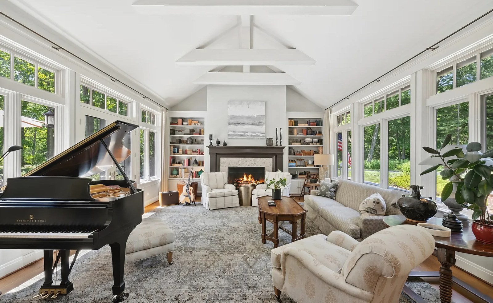 The living room under a white collar-beam ceiling, windows on three sides, with a fireplace and built-in bookcases on the end wall and a grand piano in the near corner.