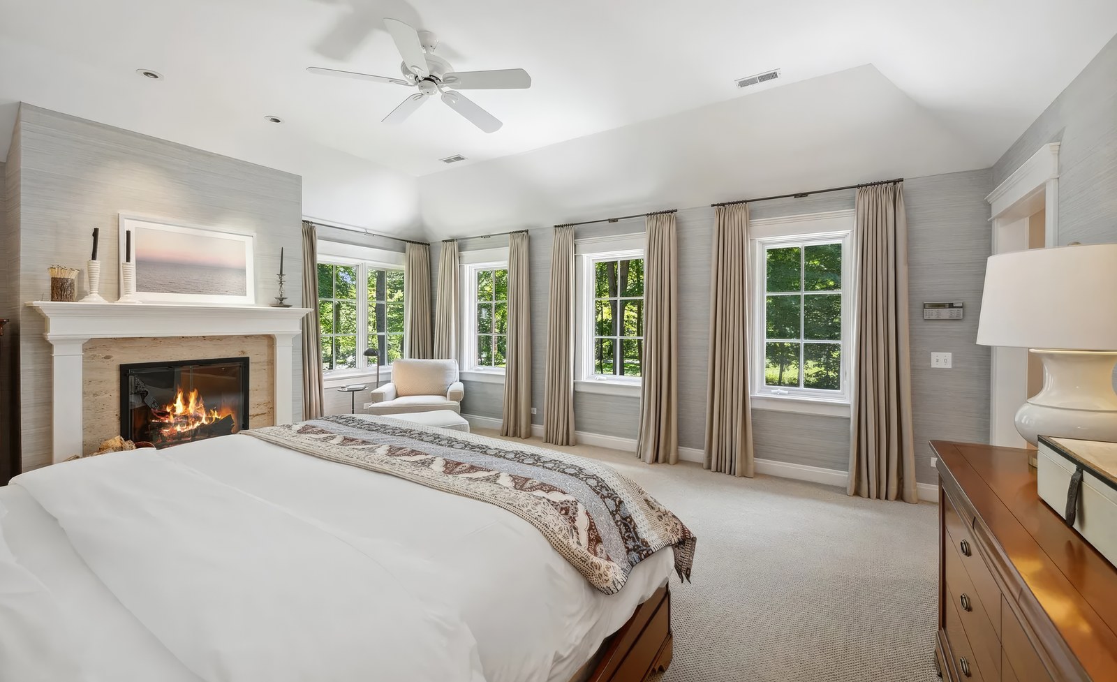 The primary bedroom, with a fireplace and mantel on the near wall and a run of windows along the far one, looking into the trees.