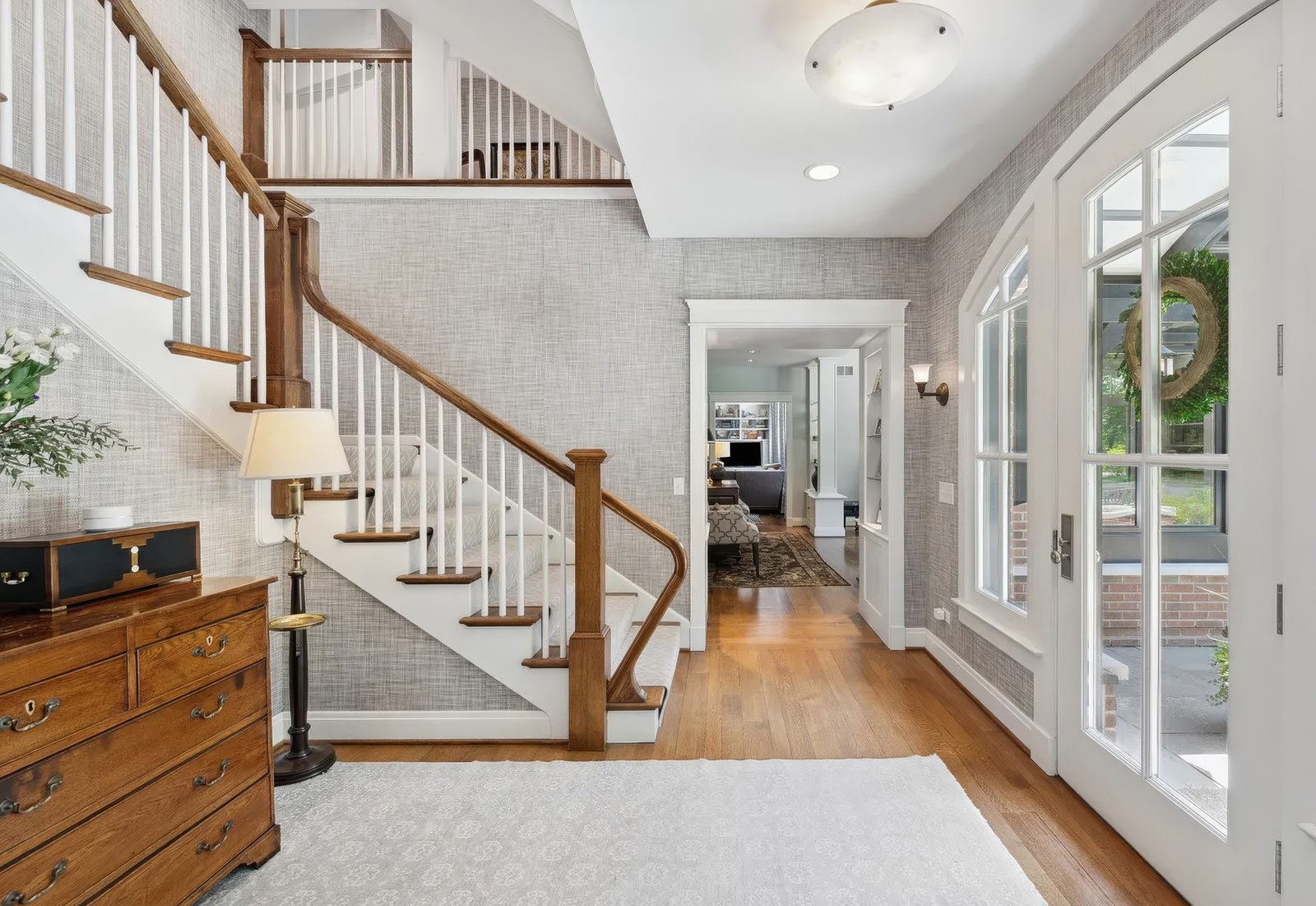 The stair hall on the entrance floor, with a turned stair in stained oak against white balusters and a doorway through to the living rooms beyond.