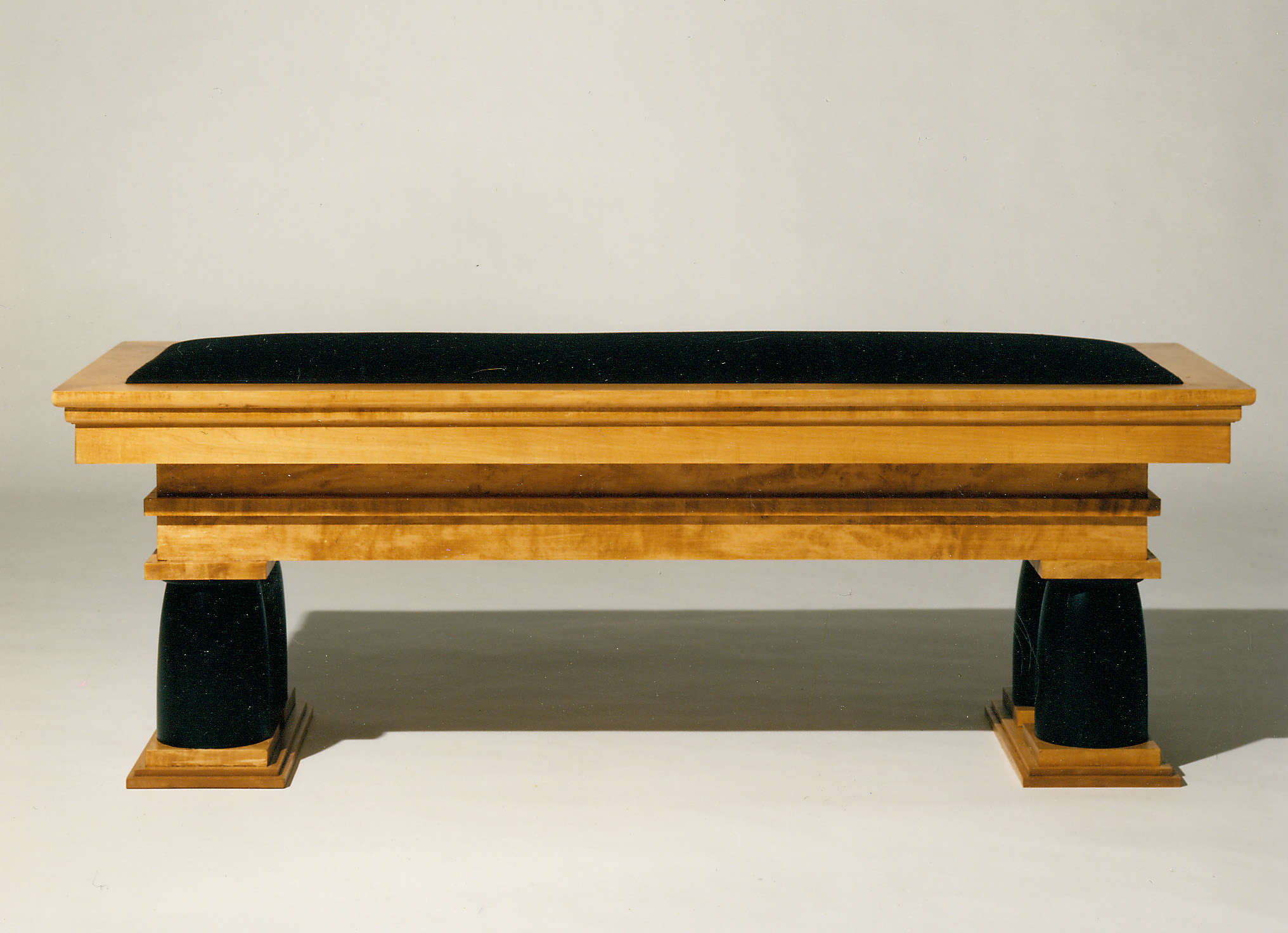 A long bench in pale birch with a black velvet seat, its frame moulded like a cornice and carried on two tapered black legs standing on stepped plinths.