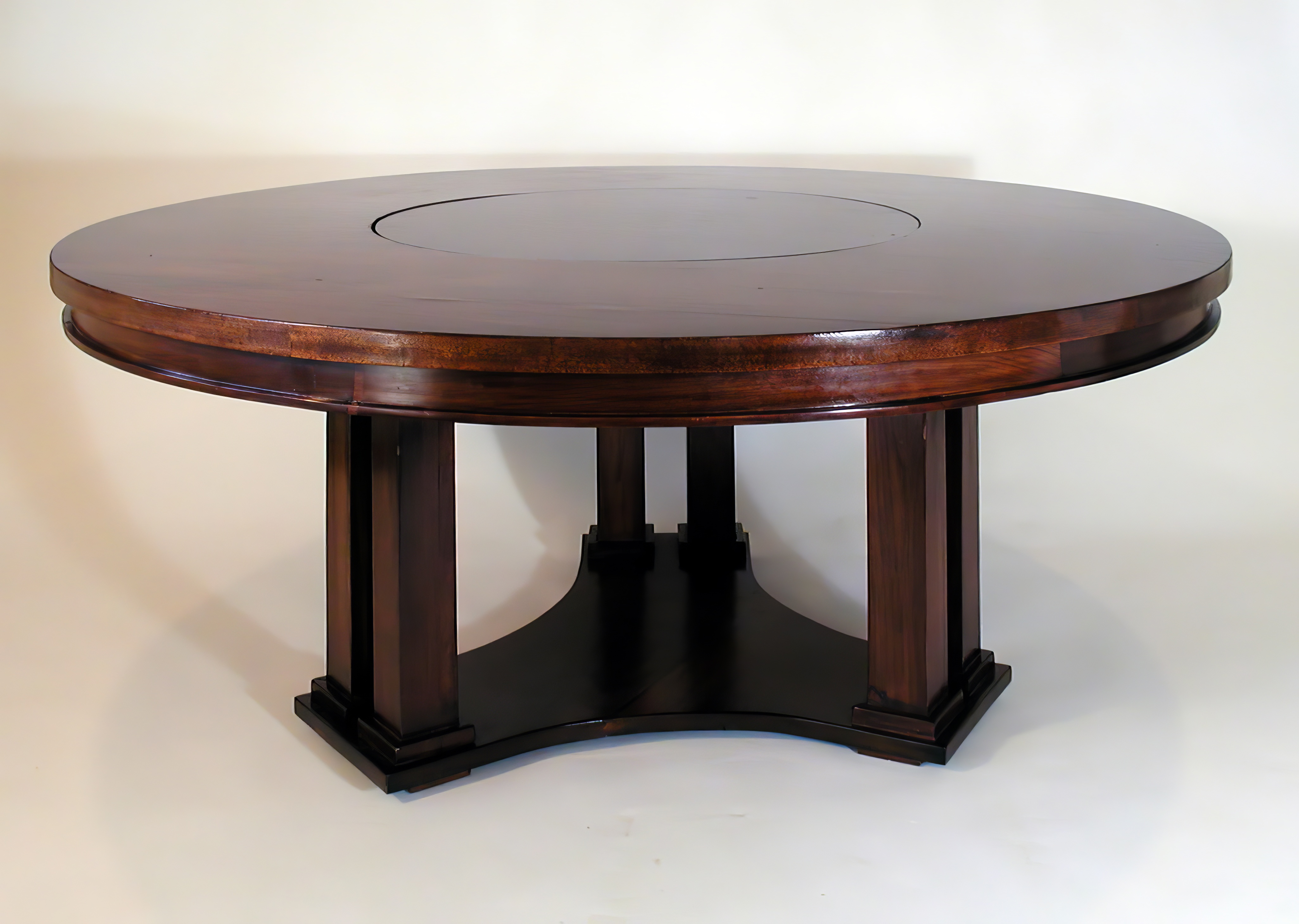 A round mahogany dining table with a burl banded edge and a revolving centre panel, standing on four dark tapered piers over a curved platform base.