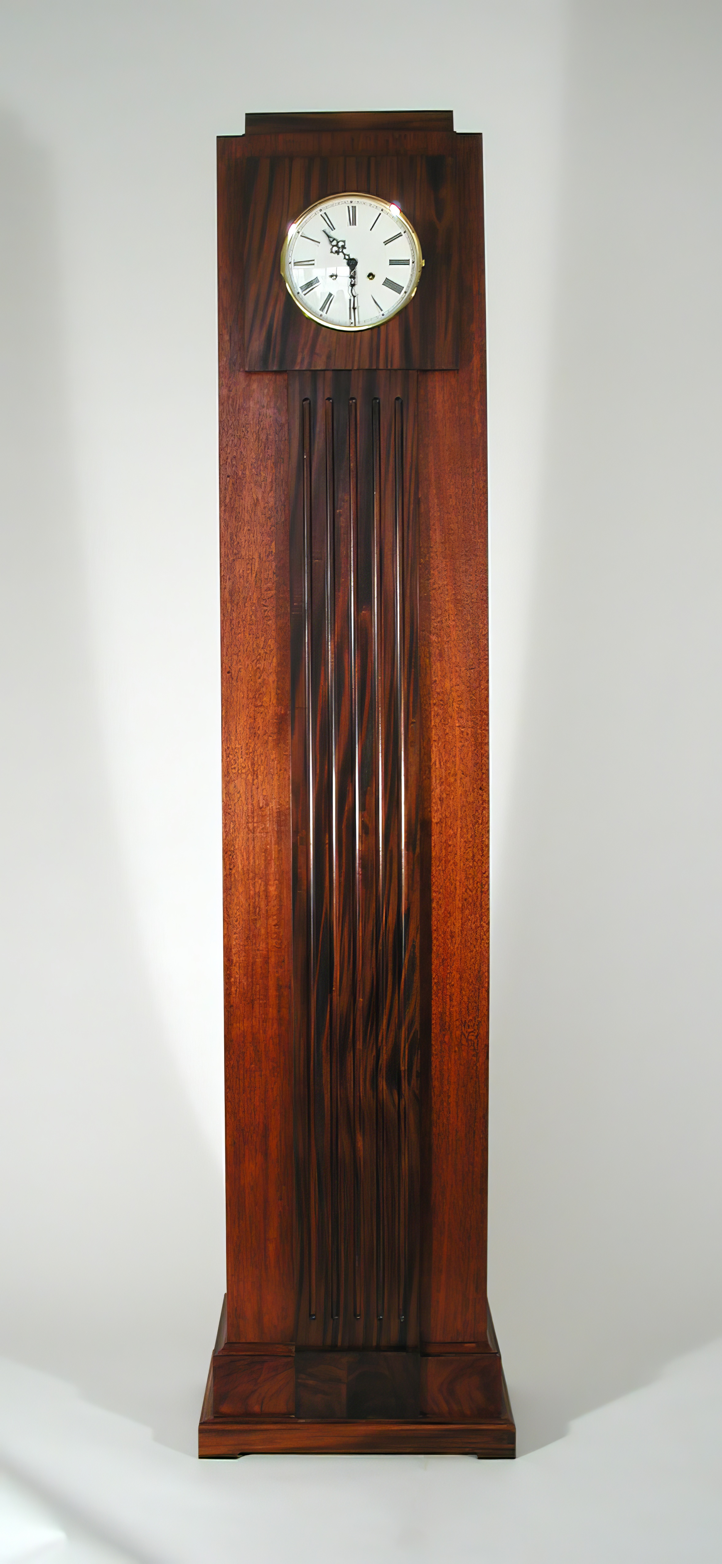 A tall case clock in mahogany, a dark figured ebony panel running the full height of the trunk beneath a white enamel dial in a brass bezel.
