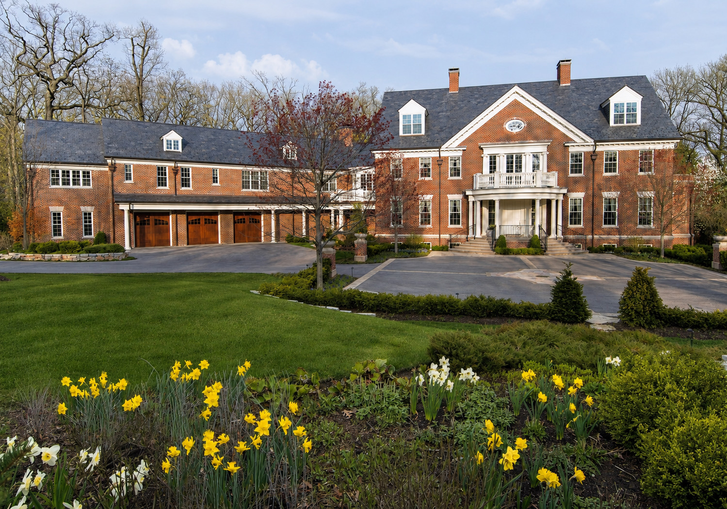 The house after the work, seen across the lawn in spring: a red brick Georgian front with a columned entrance portico carrying a balustraded balcony, dormers set into a slate roof, and the rebuilt garage wing running away to the left behind three timber doors.