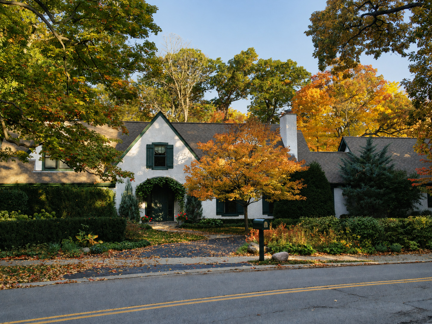 The house from the road in autumn: white stucco gables with dark green shutters set back behind clipped hedges, a maple in full colour on the lawn, and an ivy-grown arched hood over the front door.