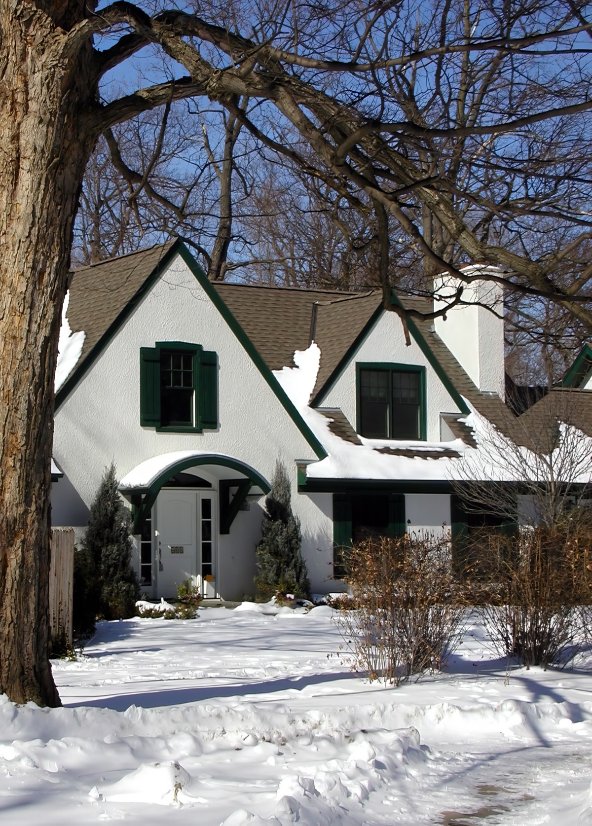 The front of the house in winter, seen past the trunk of a mature tree: white stucco gables with dark green shutters and green-edged eaves, an arched hood over the front door, and a covered porch running away to the right under a snow-covered roof.