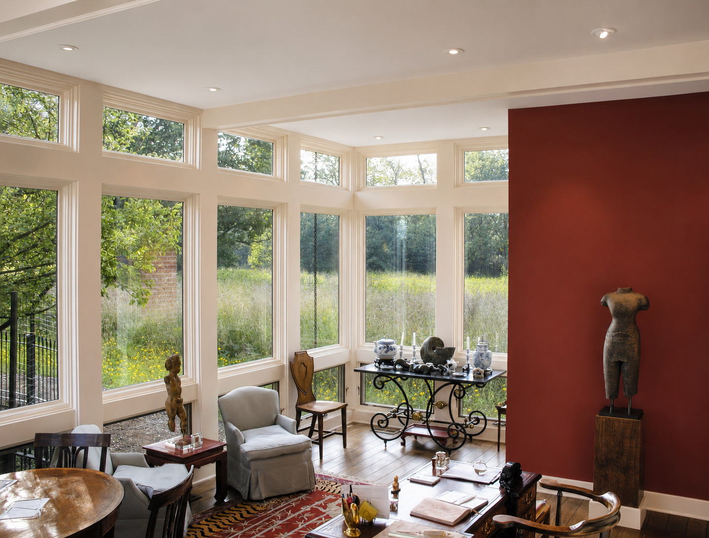 The office looking into its glazed corner, with white-framed windows running from floor to ceiling on two sides, a deep terra-cotta wall to the right carrying a sculpture on a plinth, and prairie filling every opening.