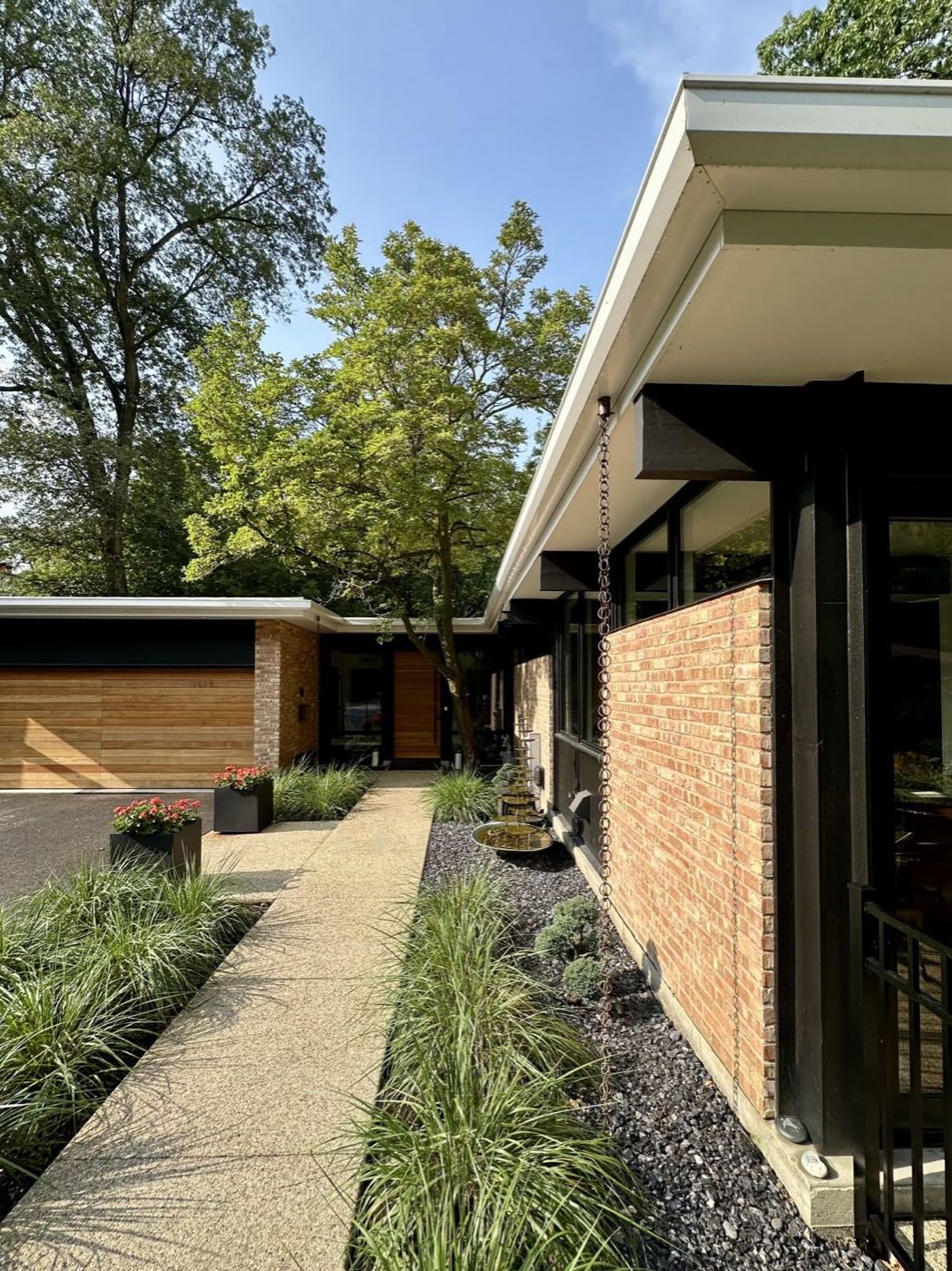 Aggregate walk running alongside the brick wall of the house toward the front door, with a rain chain and ornamental grasses.
