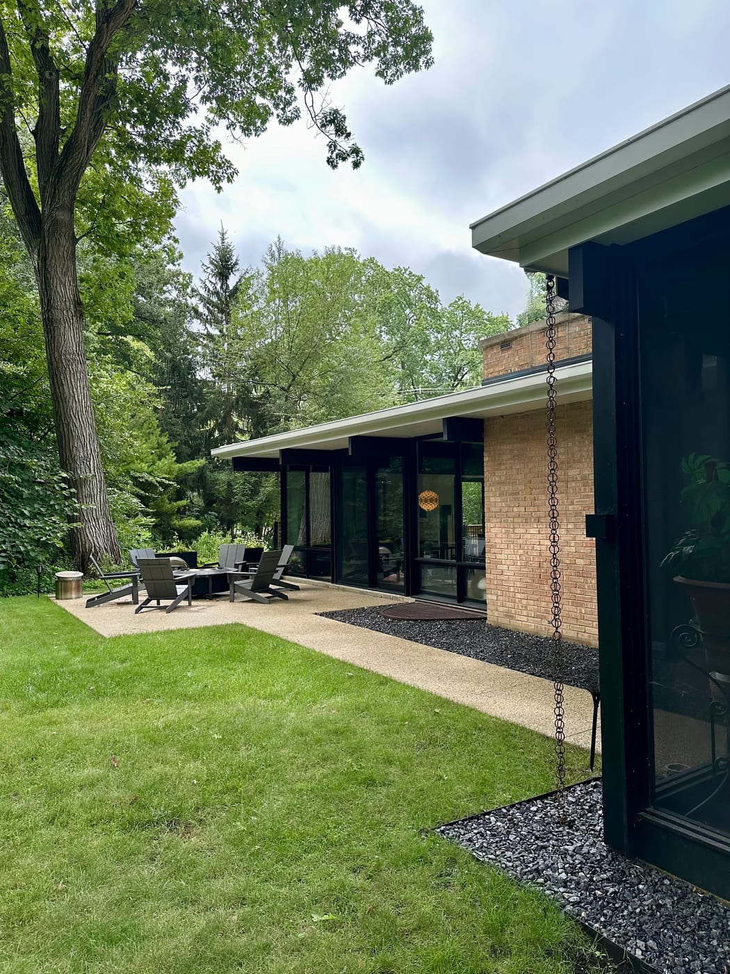 The garden elevation seen along its length from the lawn, a rain chain hanging off the eave in the foreground, the brick chimney beyond it and the terrace chairs at the far end.