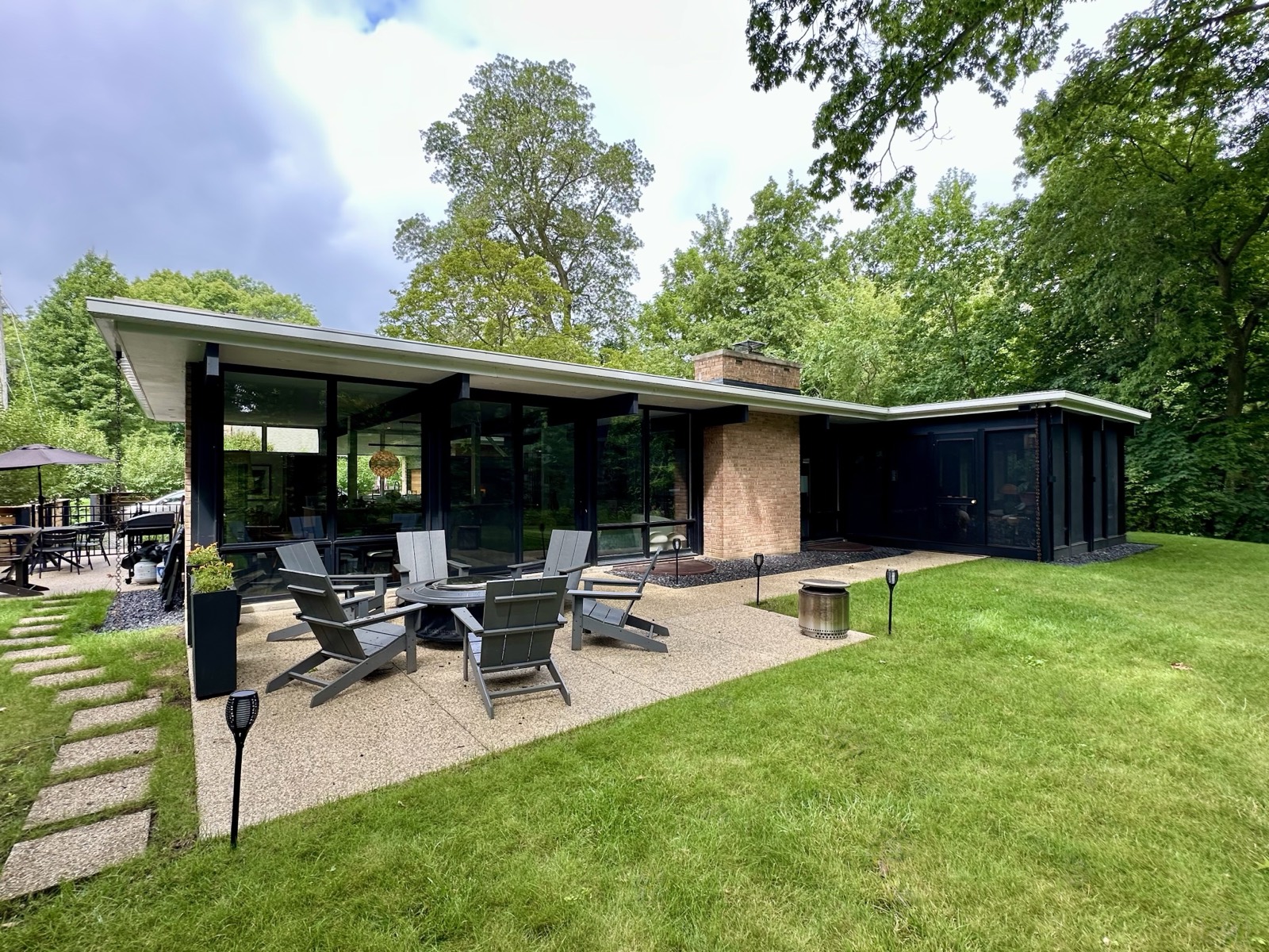 The garden side of the house across the lawn, a long low roof with a deep eave over black walls and full height glazing, a brick chimney part way along and a screened porch at the far end.