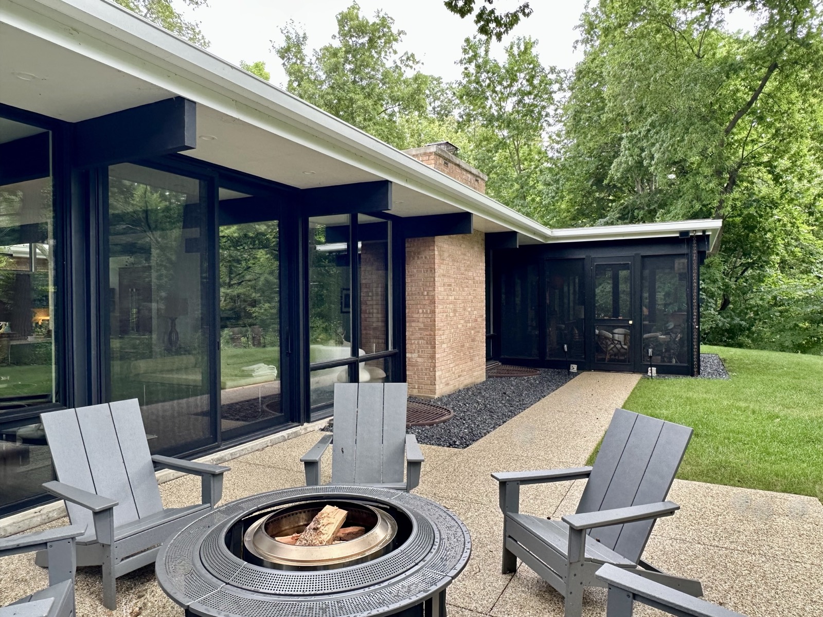 The terrace outside the living room, with a fire pit table and chairs, a glazed wall to the left and the screened porch beyond the brick chimney.