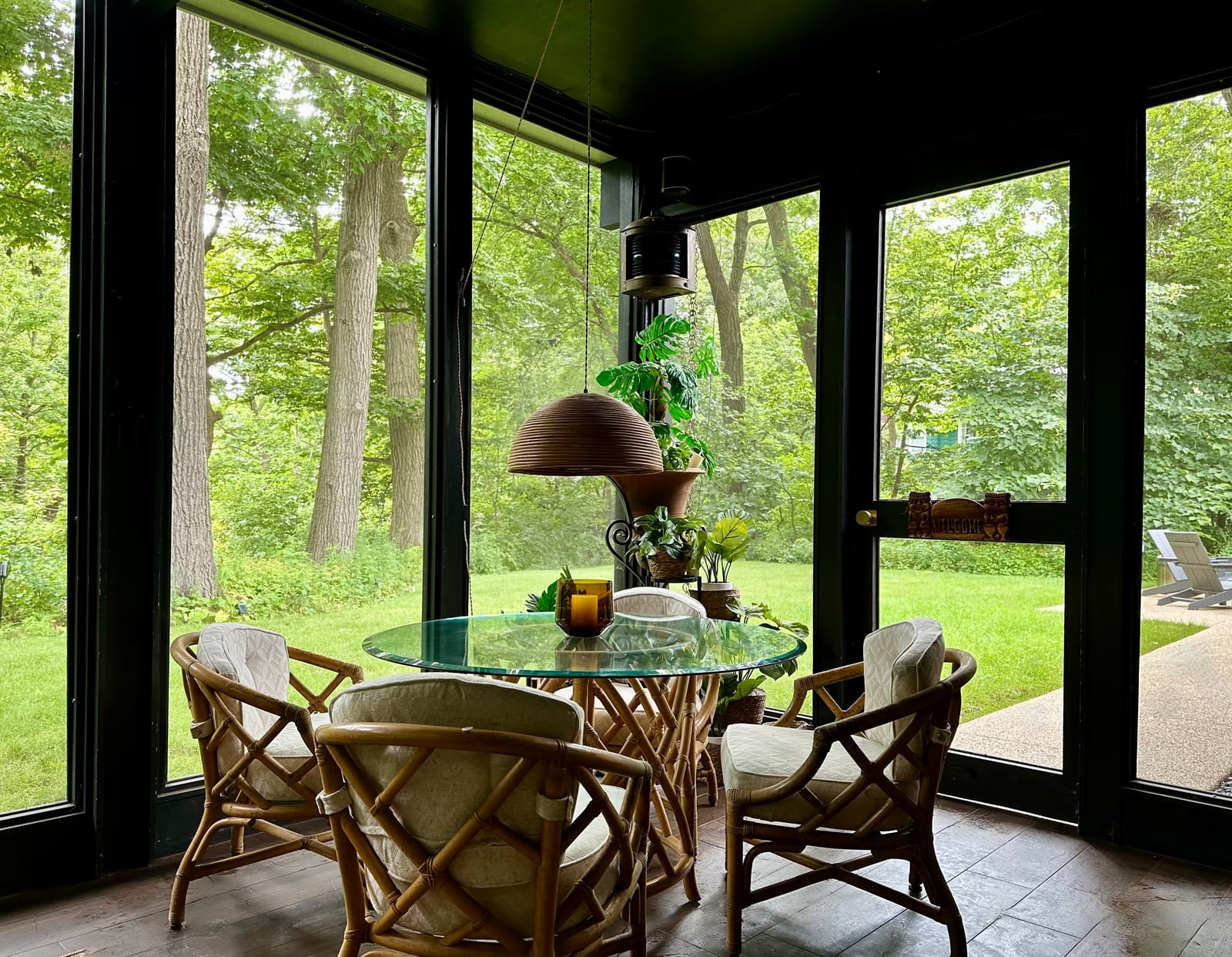 A round glass dining table and four rattan chairs under a woven pendant shade, with a screen door at the right opening onto the lawn.