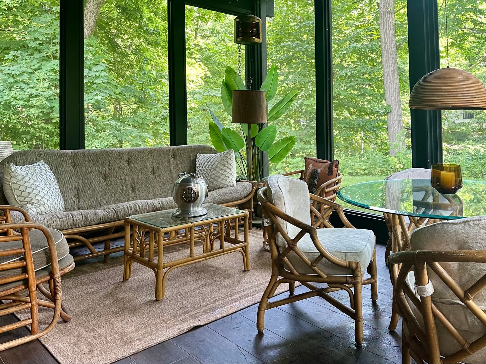 The screened porch furnished with a tufted rattan sofa, cane armchairs and a glass topped table on a jute rug, woodland filling the screens on every side.