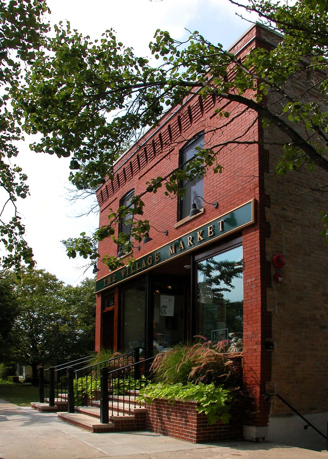 The corner entrance below a green and gold sign reading The Village Market, red brick rising past tall second floor windows to a corbelled cornice.