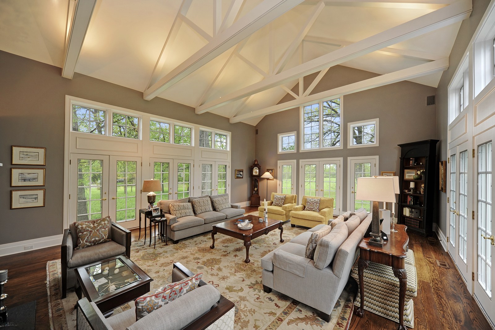 Living room under exposed white-painted roof trusses, with French doors and transom windows along both walls looking out to the meadow and oaks.